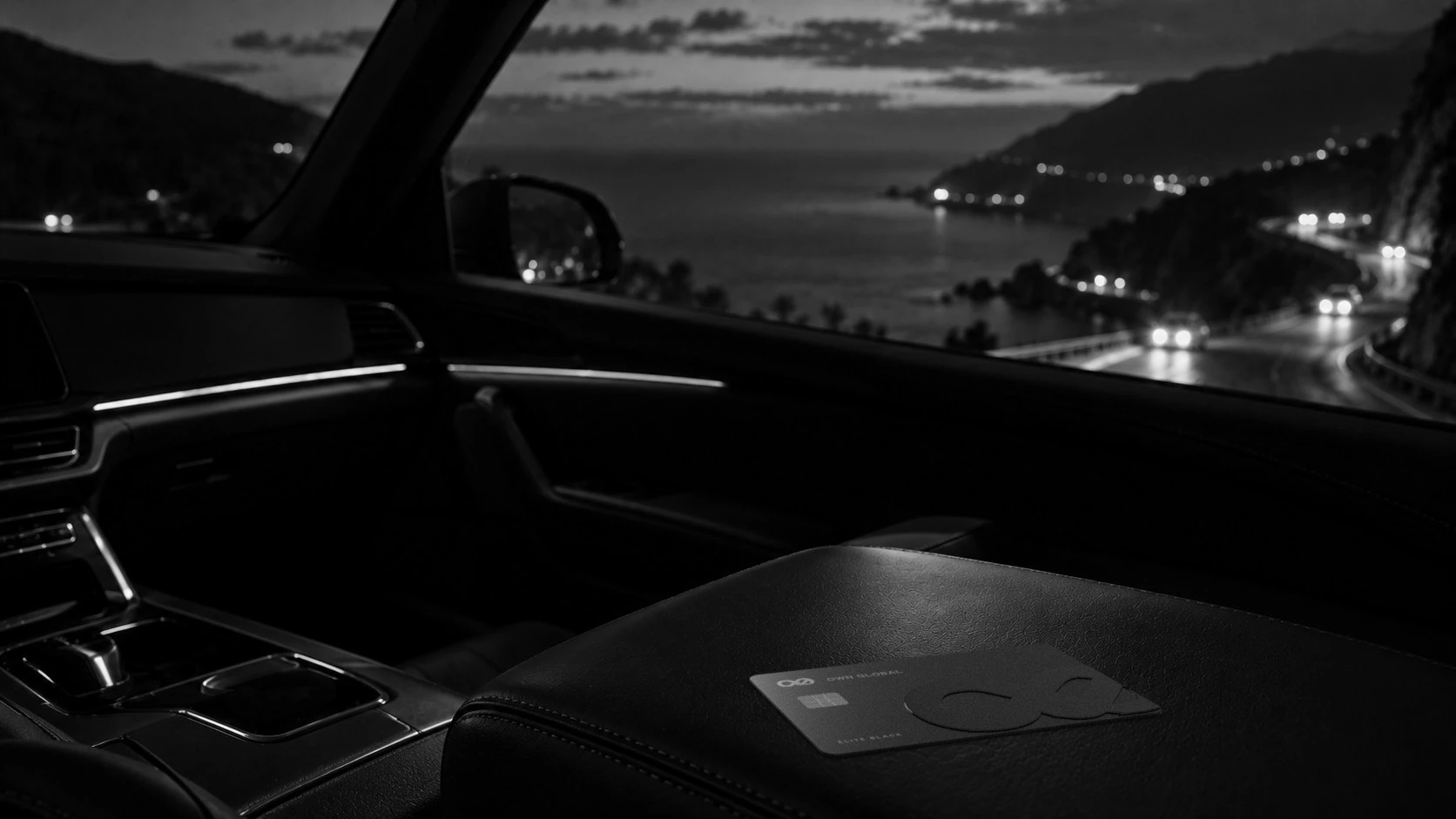 An Elite Black card on a car armrest above a coastal road at night