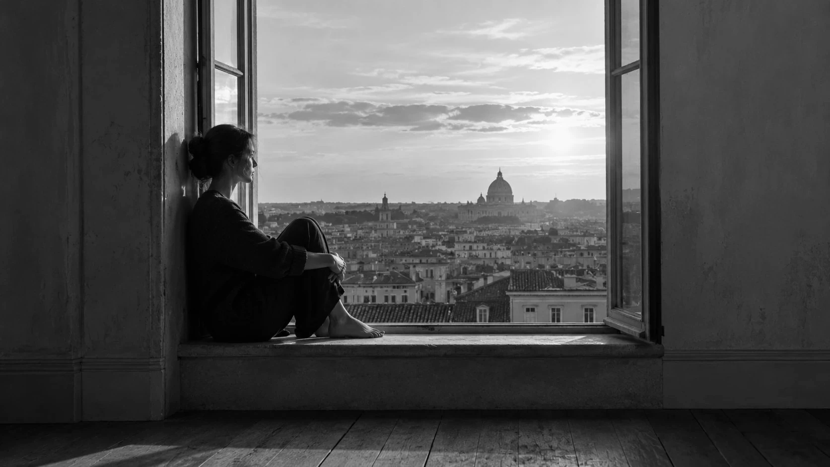 A woman resting on a window ledge above the rooftops of an old city